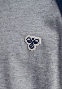 Graues Stoff-T-Shirt mit einem kleinen gestickten Bienenlogo in Marineblau und Weiß, das auf der linken Seite der Brust platziert ist.