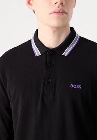 Polo shirt preto feito de tecido texturizado, apresentando um colarinho listrado contrastante em cinza e roxo, com um logotipo roxo "BOSS" no peito.