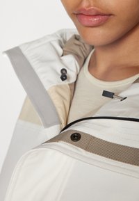 White and beige jacket with a soft texture, featuring a high collar, zippered front, and contrasting fabric accents. Hardware includes buttons and zippers.
