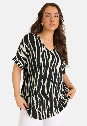 Vrouw met lang golvend haar, gouden oorbellen, een zwart-wit zebraprint V-hals top, witte broek en gouden armbanden tegen een effen achtergrond.