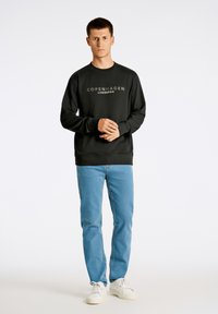 Sudadera negra con "Copenhague Lindbergh" impreso en blanco, combinada con jeans azules y zapatillas blancas. Diseño y ajuste simples.