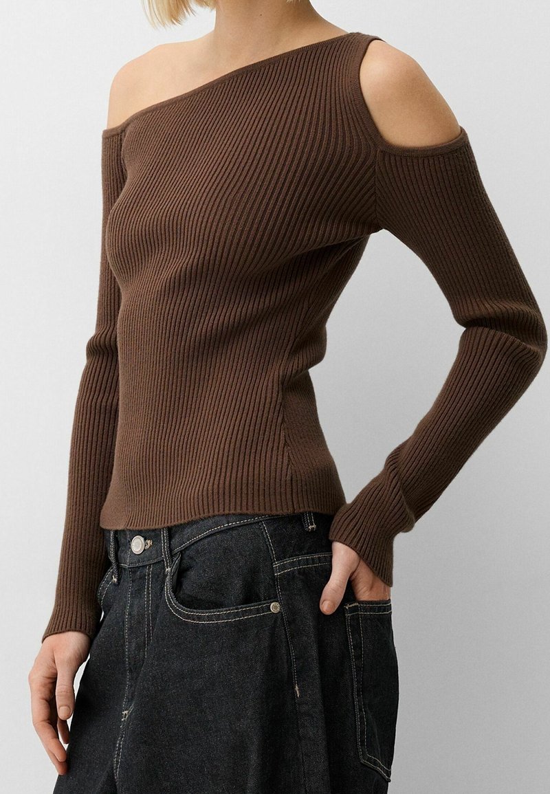 Femme portant un pull en maille côtelée marron avec épaule froide asymétrique et manches longues, associé à un jean en denim noir.