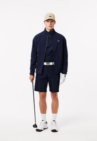 Chaqueta navy con cremallera, pantalones cortos a juego y camiseta polo; gorra beige y guante de golf blanco; sosteniendo un palo de golf; calzado deportivo.