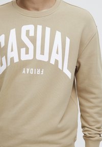 Béžová mikina s velkým bílým nápisem "CASUAL FRIDAY". Vyrobena z měkké tkaniny, má kulatý výstřih a žebrované manžety.