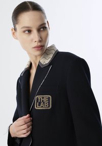Zwarte blazer met een contrasterende zigzagpatroon kraag, een gouden geborduurd embleem met de tekst "THE LAB" en zichtbare witte stiksels.