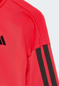 Camiseta deportiva roja con un tejido texturizado, que presenta acentos de tres rayas negras en cada hombro. Diseñada para ofrecer un aspecto ajustado.
