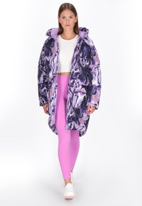Pufferjack met een paarse en zwarte marmerprint, met een capuchon, een ritssluiting aan de voorkant en twee grote zakken. Draagt het met roze legging en witte schoenen.