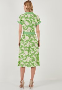 Blumenkleid aus lebhaft grünem Stoff mit weißem Blumenmuster, kurzen Ärmeln, tailliertem Gürtel und knielanger Silhouette. Schlicht grüner Kragen und Bündchen.