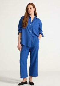 Modrá lněná košile na knoflíky s náprsní kapsou. V kombinaci s volnými kalhotami ve stejném odstínu. Černé ploché boty doplňují celý outfit.