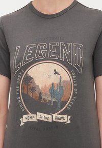 Tamsiai pilkas marškinėliai su apvaliu grafiniu vaizdu, kuriame yra kaktusų ir kalnų, tekstas "LEGEND" ir "HOME OF THE BRAVE" stilizuoti kreminės ir mėlynos spalvos.