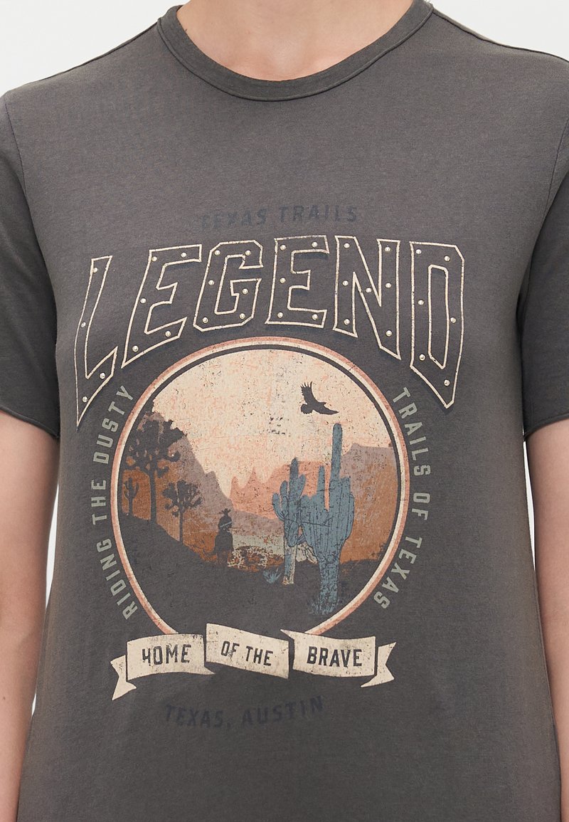Camiseta gris oscuro con un gráfico circular que presenta cactus y montañas, con el texto "LEGENDA" y "HOGAR DE LOS VALIENTES" estilizado en crema y azul.