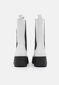 Botas de tornozelo em preto e branco com um design elegante, feitas de material brilhante, apresentando laterais elásticas e uma sola grossa preta.