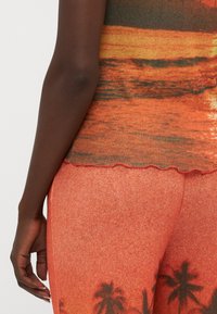 Top de tirantes multicolor con un diseño de atardecer, tejido texturizado y dobladillo con forma de concha. Combinado con pantalones naranja ajustados y texturizados que presentan motivos de palmeras.