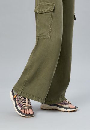 Pies con sandalias deportivas de tiras abiertas y con cordones, en tonos púrpura y beige, combinadas con pantalones cargo anchos de color verde oliva.