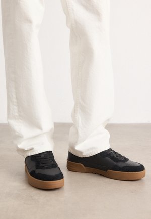 Person iført hvide bukser og sorte sneakers med brune såler, stående på en glat lysegrå gulv.