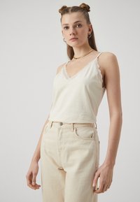 Ljusbeige linne med spetsdetaljer vid V-ringningen. Tyget ser mjukt och lätt ut, matchat med krämfärgade högmidjade jeans.
