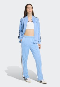 Woman standing wearing light blue Adidas tracksuit with black and white stripes, white crop top, and white sneakers with black stripes.