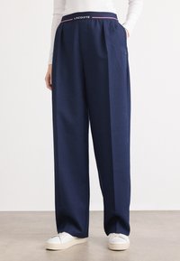 I pantaloni a gamba larga blu navy presentano un design plissettato, una vita elasticizzata con il logo "LACOSTE" e sono realizzati in un tessuto morbido e leggero.