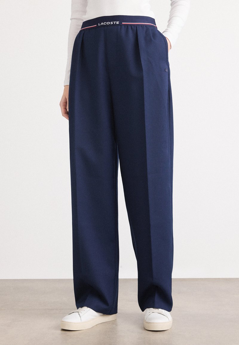 I pantaloni a gamba larga blu navy presentano un design plissettato, una vita elasticizzata con il logo "LACOSTE" e sono realizzati in un tessuto morbido e leggero.