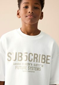 Vit bomullströja med stora metalliska gulda bokstäver som visar "SUB5CRIBE", geografiska koordinater och "FUTURE SYSTEMS" i olika storlekar.