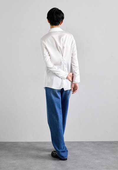 Camicia bianca in satin a maniche lunghe con un dettaglio di bottone sul retro, abbinata a pantaloni in denim blu larghi e scarpe nere, vista da dietro.