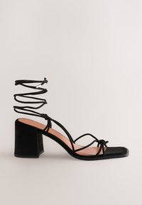 Sandalo con tacco in suede nero con punta quadrata, sottili cinturini incrociati e cinturino alla caviglia a spirale. Presenta un tacco largo per maggiore supporto.