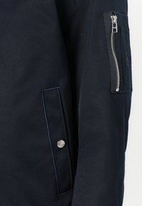 Veste bleu marine au toucher lisse, dotée d'une poche zippée sur la manche, de coutures marquées et d'un bouton pression décoratif.