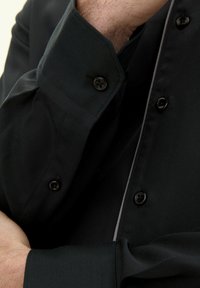 Camicetta nera con colletto strutturato, polsini con bottoni e una sottile piping in contrasto lungo la parte anteriore. Tessuto dalla trama liscia.