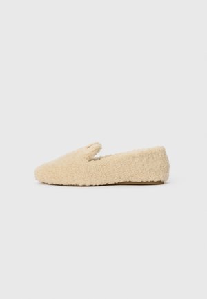Cremefarvet fluffy slip-on sko med lav hæl og blødt, tekstureret stof, vist i siden på en hvid baggrund.