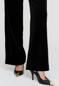 Schwarze Samt-Hosen mit weitem Bein, kombiniert mit spitzen schwarzen High Heels, die eine goldene Zehenakzentuierung haben. Die Schuhe verfügen über einen schlanken Absatz und ein elegantes Design.