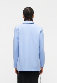 Άτομο με πλεξούδες, φορώντας γαλάζιο σακάκι και μαύρη φούστα, στέκεται με την πλάτη στην κάμερα μπροστά σε λευκό φόντο.