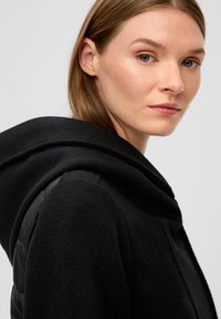 Manteau noir avec une grande capuche, présentant un tissu texturé et des sections rembourrées sur les épaules. Design simple sans matériel visible.