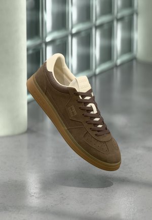 Brauner Wildledersneaker mit Gummisohle und dunkelbraunen Schnürsenkeln, schwebend über einem grauen, polierten Boden und einer Wand aus Glasbausteinen.