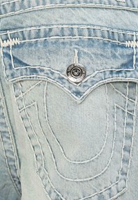 Un bolsillo de mezclilla azul claro con costuras blancas, un botón de metal plateado con logo y un diseño de costura curvada distintivo.