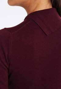 Pull en tricot bordeaux avec un col à revers, présentant une texture lisse et une silhouette ajustée. Détail des coutures aux épaules visible.