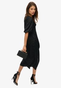 Vestido negro midi con mangas abullonadas, tejido texturizado y silueta ajustada. Combinado con tacones de aguja negros con tiras y un clutch de mimbre.