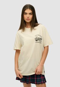 Camiseta de manga corta beige con un logo negro impreso en el lado izquierdo del pecho, combinada con una falda de cuadros con pliegues en tonos azul marino, rojo y verde.