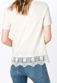 Top corto de manga corta en color crema con un dobladillo de encaje con patrones circulares; combinado con jeans de color azul claro, mostrando un estilo casual.