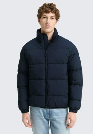 Ung mand med krøllet hår iført en lynlås mørkeblå puffjakke og lyseblå jeans, stående mod en ensfarvet baggrund.