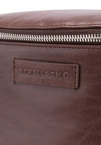 Braune Ledertasche mit strukturierter Oberfläche, ausgestattet mit einem silbernen Reißverschluss und einem geprägten "Strellson"-Logo auf einem rechteckigen Lederetikett.