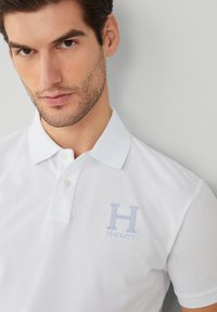 Muž s tmavými vlasy a strništěm, oblečený v bílém polo tričku se dvěma knoflíky a modrým logem "H Hackett" na hrudi, na světle šedém pozadí.