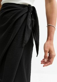 Jupe portefeuille noire en tissu texturé, dotée d'un lien noué. Gros plan sur la main droite tenant la jupe avec un bracelet en argent au poignet.