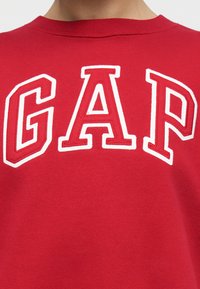Κόκκινο φούτερ με μεγάλα λευκά γράμματα με ανάγλυφη περίγραμμα που σχηματίζουν τη λέξη "GAP" στο πάνω μέρος της πλάτης, κάτω από τη γραμμή του λαιμού.