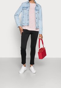 Giacca di denim chiaro, maglietta a righe rosse e bianche, pantaloni neri e sneakers bianche. Tiene una borsa strutturata rossa. Texture morbide e stile casual.