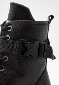 Stivale alla caviglia in pelle nera con superficie texturizzata, progettato con lacci, una fascia in tessuto con fibbia e dettaglio del logo sul lato.