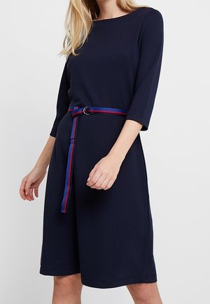 Vrouw draagt een marineblauwe jurk tot op de knie met driekwartmouwen en een dunne gestreepte blauw-rode riem in de taille.