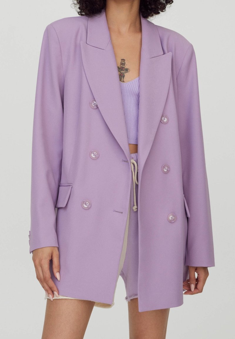Personne portant un blazer croisé violet clair sur un crop top assorti et un short beige à cordon. Tatouage visible sur la poitrine.