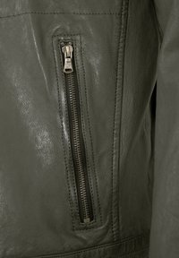 Olivgrüne Lederjacke mit einer vertikalen Reißverschlusstasche mit Metallic-Reißverschluss und kontrastierenden Nähten. Weiche, strukturierte Oberfläche.