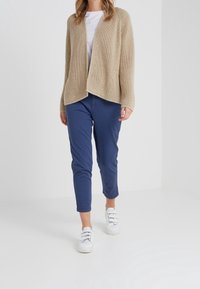 Beige pletený cardigan s volným předním dílem, spárovaný s námořnickými kalhotami a bílými teniskami. Tkanina má texturovaný, otevřený vzor.