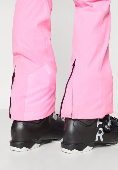 Svetlo roza smučarske hlače z zadrgami na dnu, kombinirane s črnimi smučarskimi čevlji z belimi poudarki, vidni od strani.